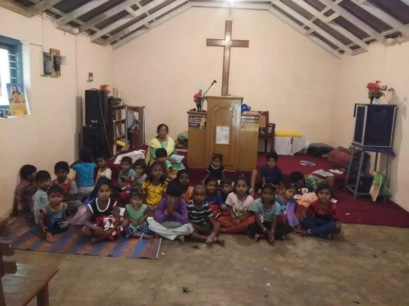 A group of children and adults seated in a room with a cross